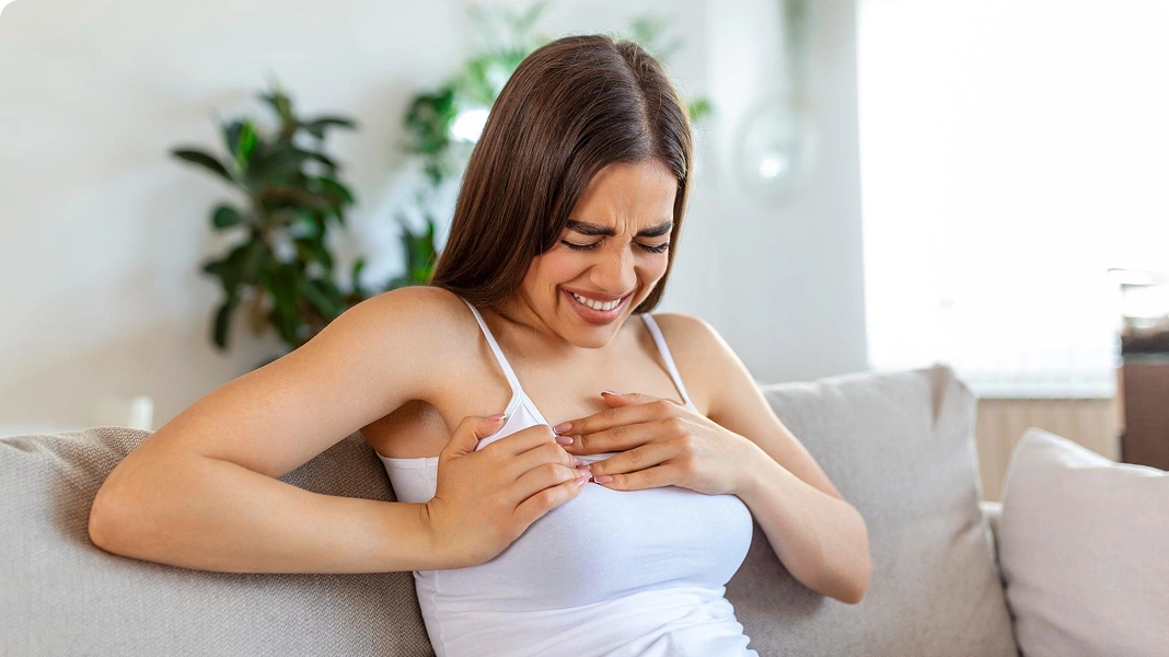 Woman in white tank top sitting on a couch, grimacing and clutching her right breast, indicating breast pain or discomfort.