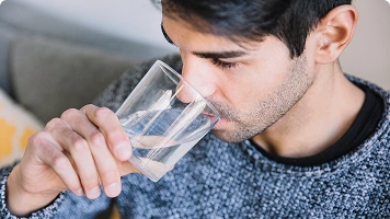 A man in a gray sweater drinking a glass of water, possibly representing hydration or increased thirst.