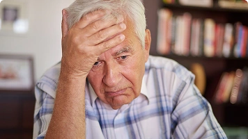 An older man in a blue shirt holding the back of his head, appearing confused or concerned