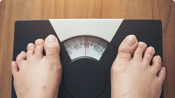 A person standing on a digital scale with a yellow measuring tape in the foreground, representing weight measurement or weight loss.